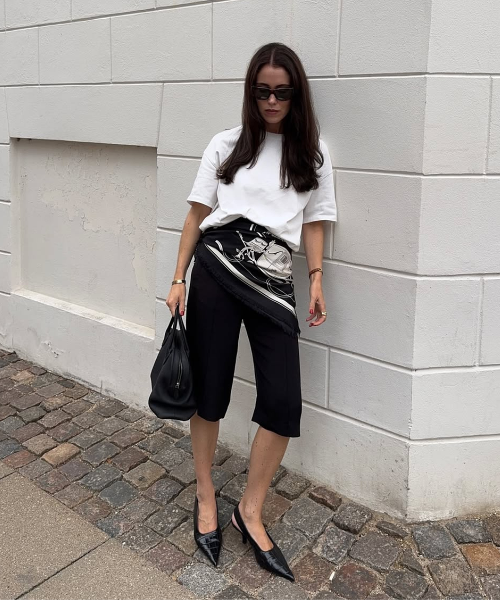 Mulher em look primavera com camiseta branca e bermuda preta, usando sapatos e bolsa pretos.