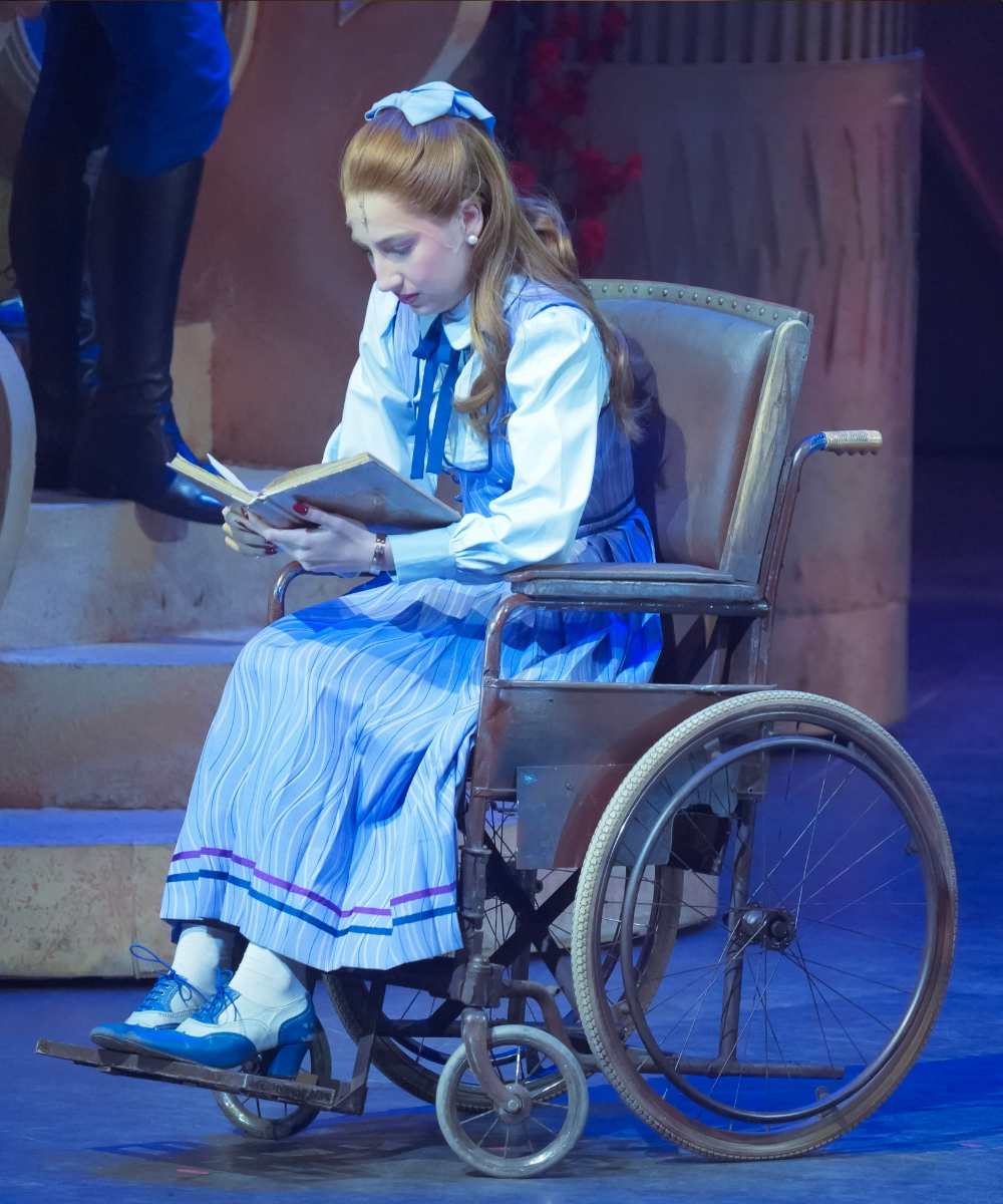 Pessoa em cadeira de rodas vestida como Nessarose lê um livro em cena teatral, criando um ambiente místico e wicked brasil