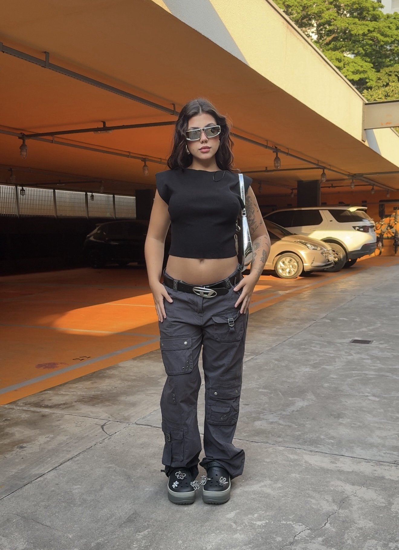 Mulher usa blusa preta, calça cargo cinza e looks estilosos com Crocs com enfeites, em um estacionamento coberto.