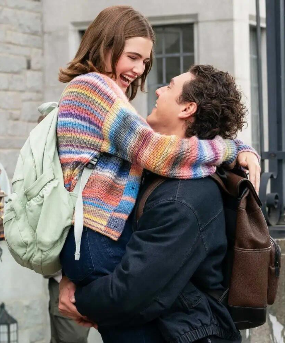 Casal se abraça e sorri em frente a um prédio. Mochilas nas costas. Cena lembra livros que viram adaptações.