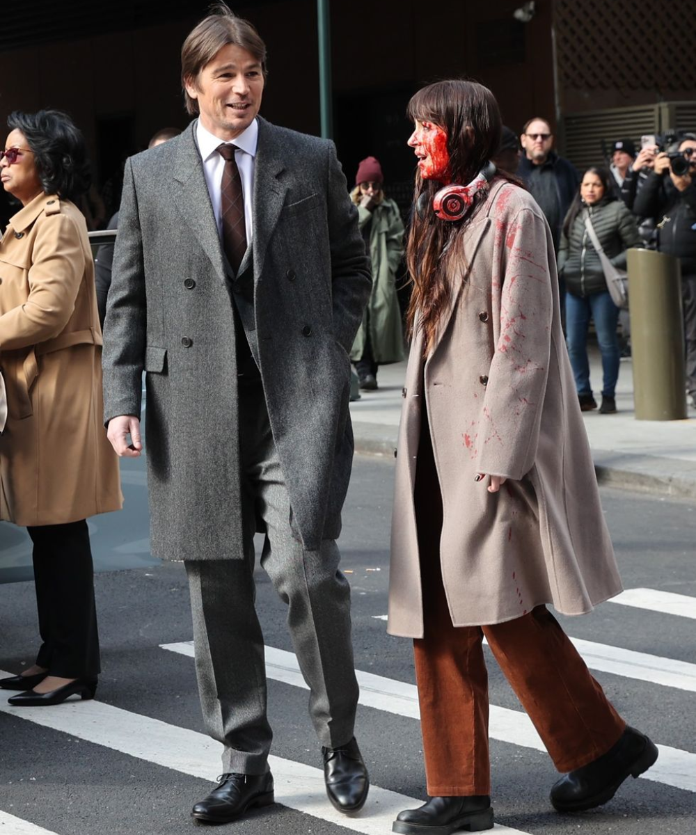 Duas pessoas caminhando na rua; uma está com o rosto manchado de vermelho, possivelmente em set de uma adaptação de livro para televisão.