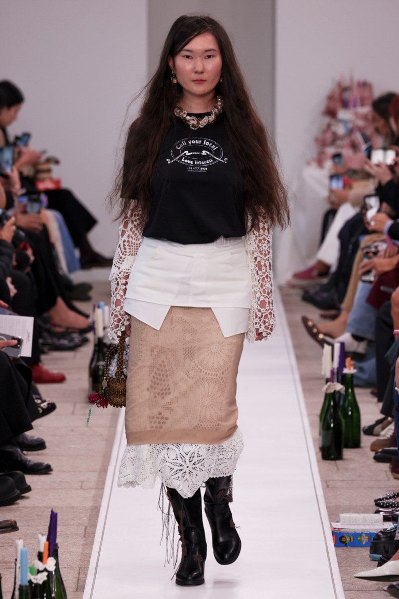 Modelo com camiseta preta e saia rendada, desfilando na Milan Fashion Week SS26. Público ao fundo.
