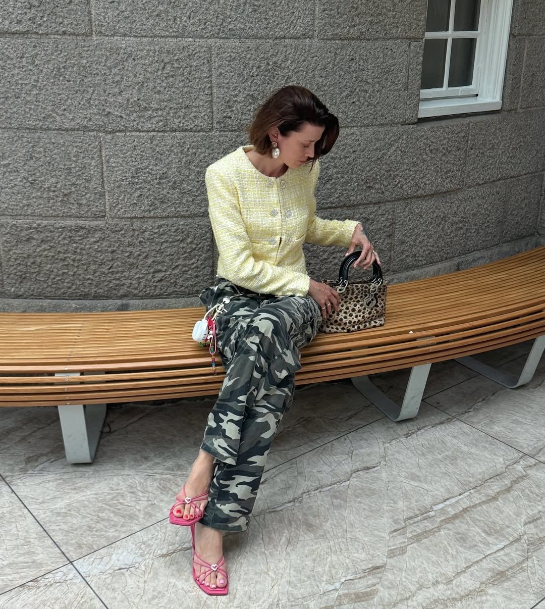 Mulher sentada em banco, usando calças com estampa camuflada e bolsa de oncinha.