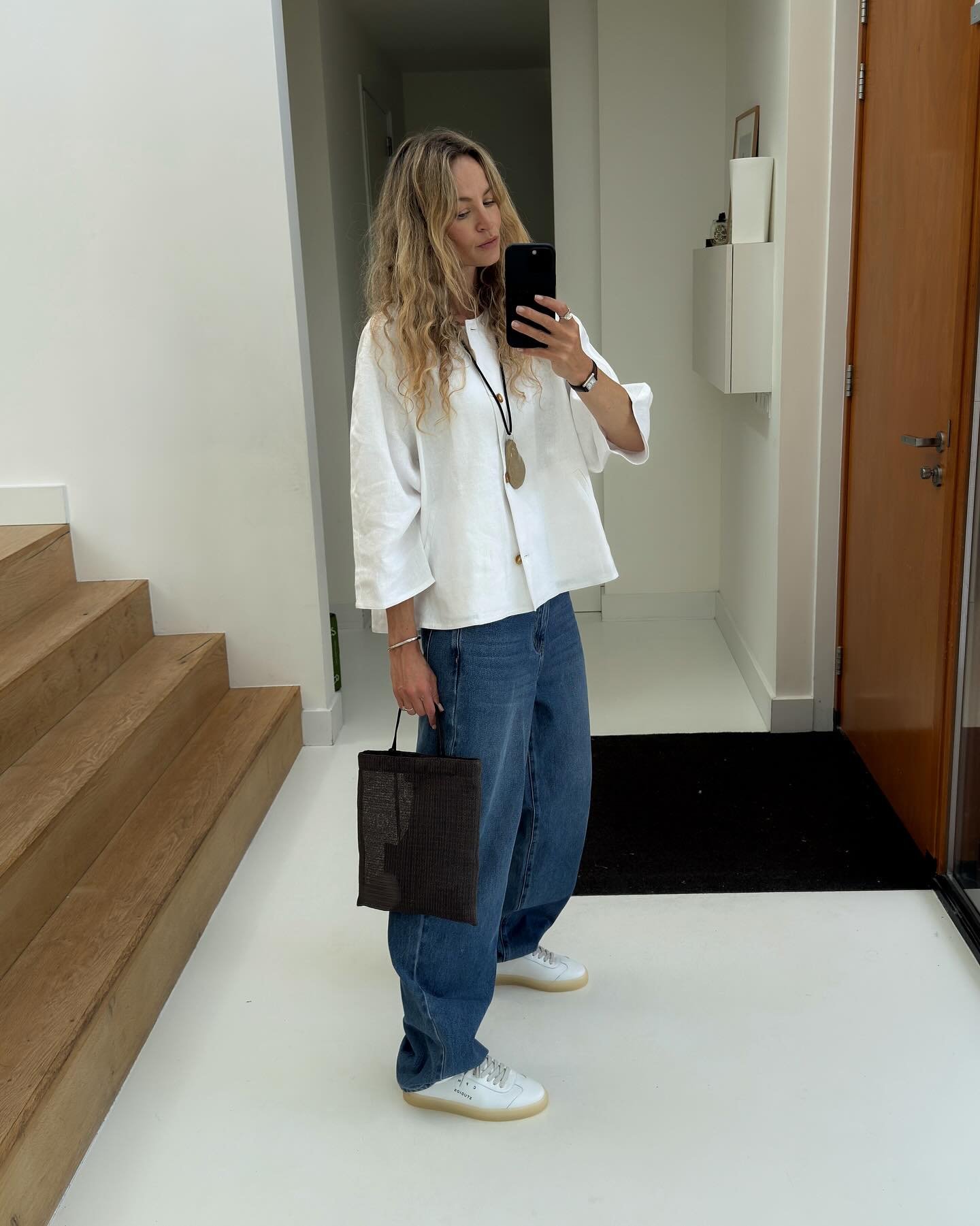 Pessoa em estilo casual de trabalho, com camisa branca, jeans e bolsa preta, em um ambiente interno minimalista.