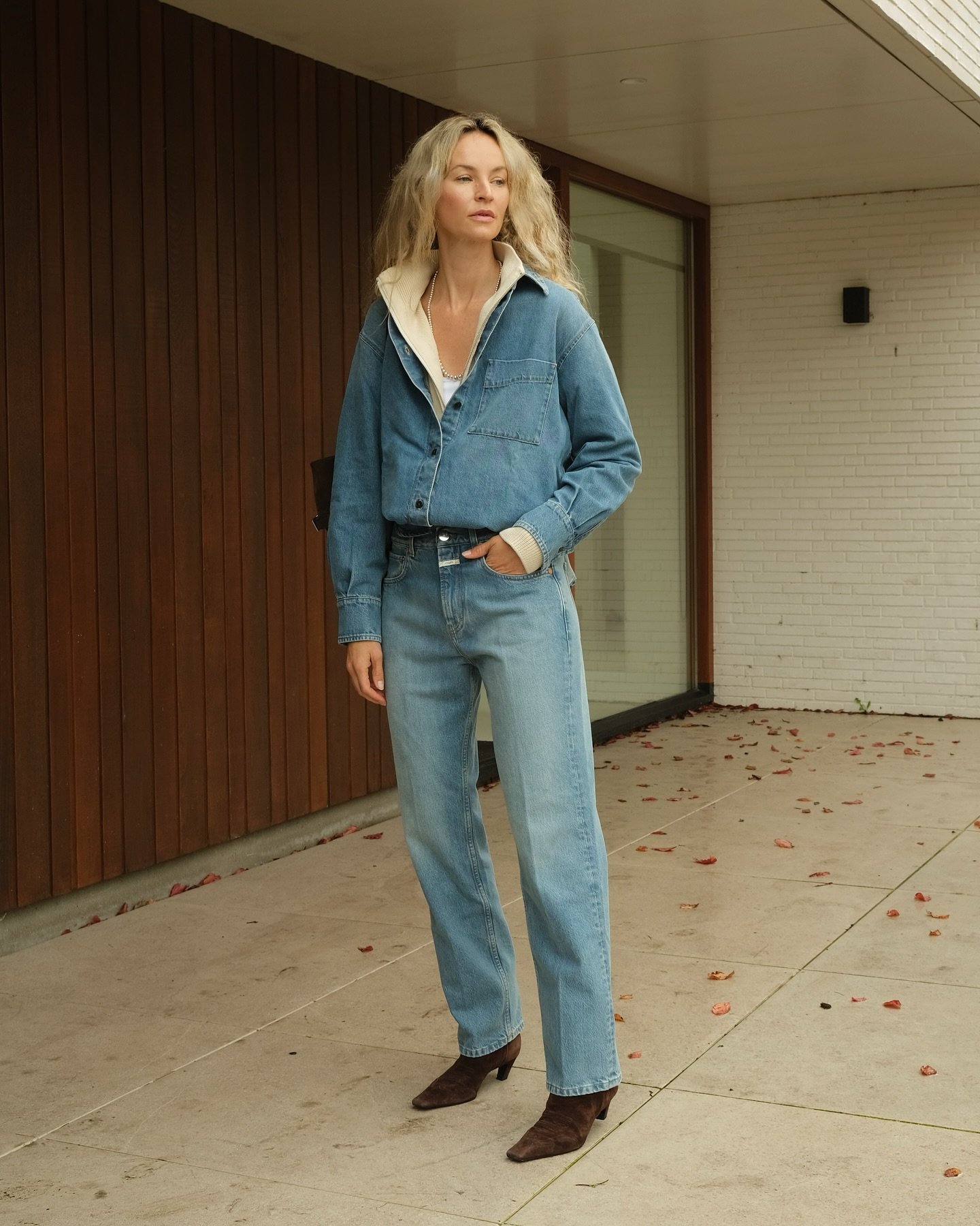 Pessoa com cabelo solto loiro vestindo jaqueta e calça jeans, em um ambiente externo. Estilo casual.