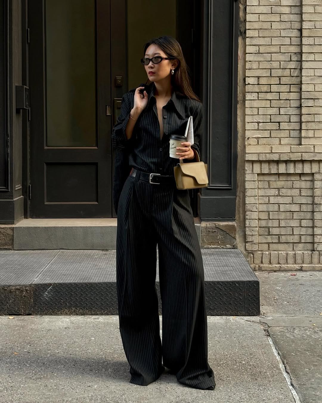 Pessoa de óculos em look formal preto com xícara de café e bolsa bege, destacando melhores looks de trabalho.