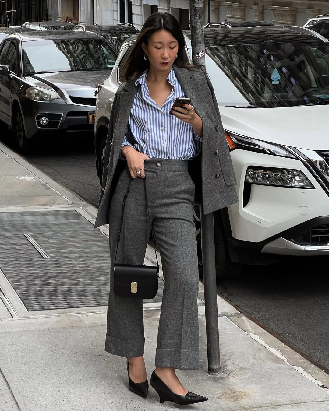 Jovem com blazer cinza sobre camisa listrada, calça social e bolsa preta, em estilo urbano elegante.