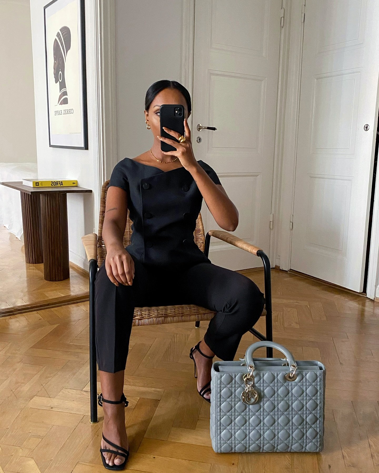 Uma mulher em traje de trabalho elegante, sentada, veste roupa preta com sandálias de salto e bolsa cinza ao lado.