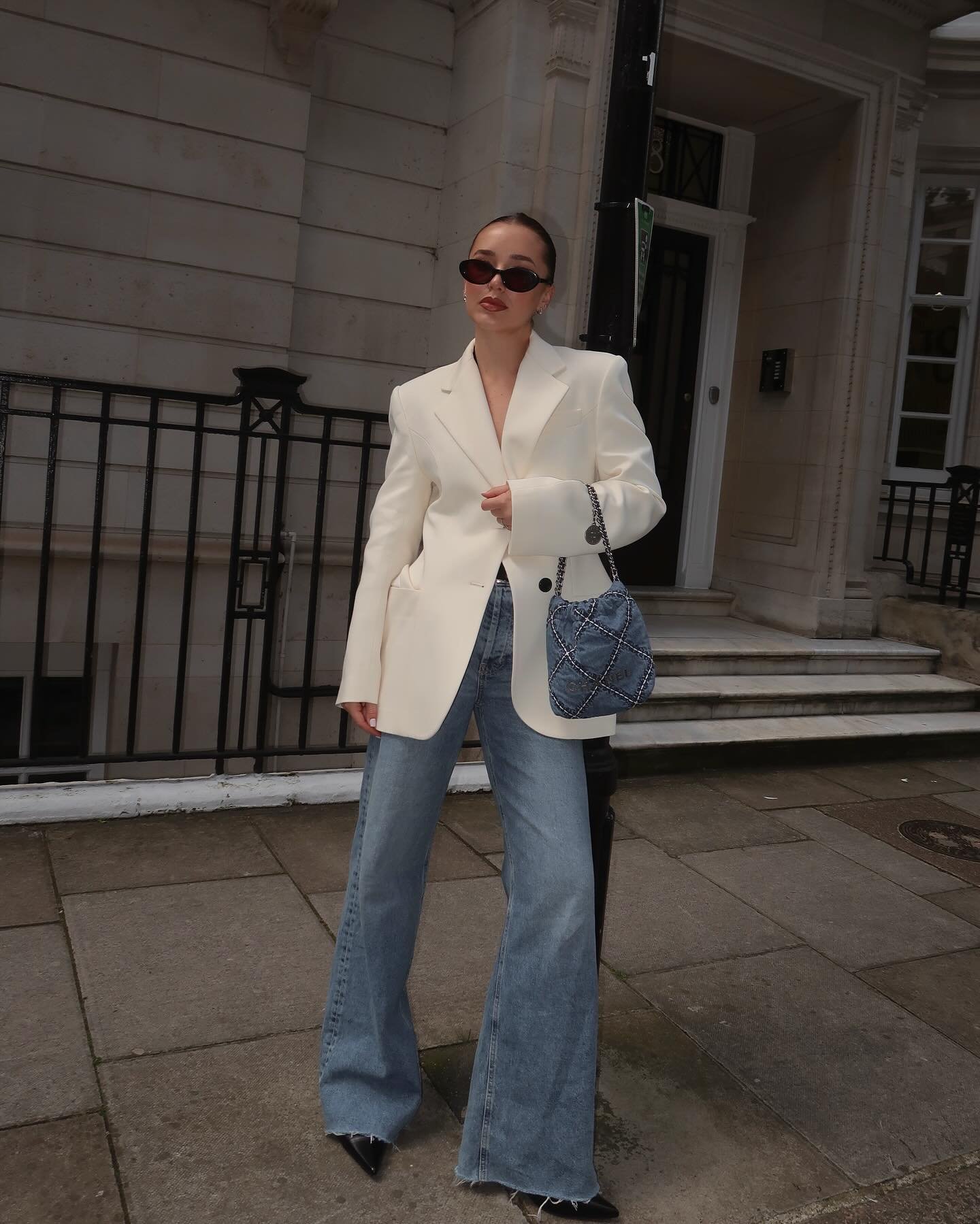 Pessoa com blazer branco e jeans flare, segurando bolsa quadriculada. Óculos escuros complementam o visual de trabalho.