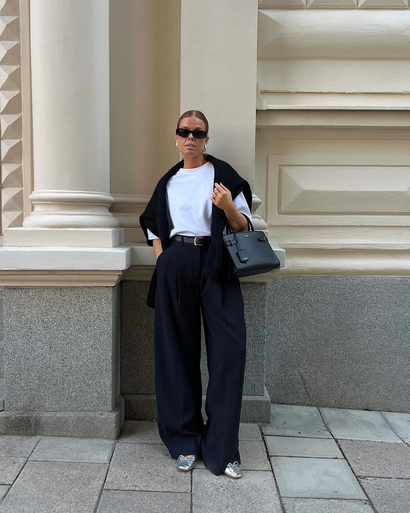 Pessoa vestindo roupas elegantes de trabalho: calça preta ampla, camiseta branca, bolsa preta, óculos de sol.