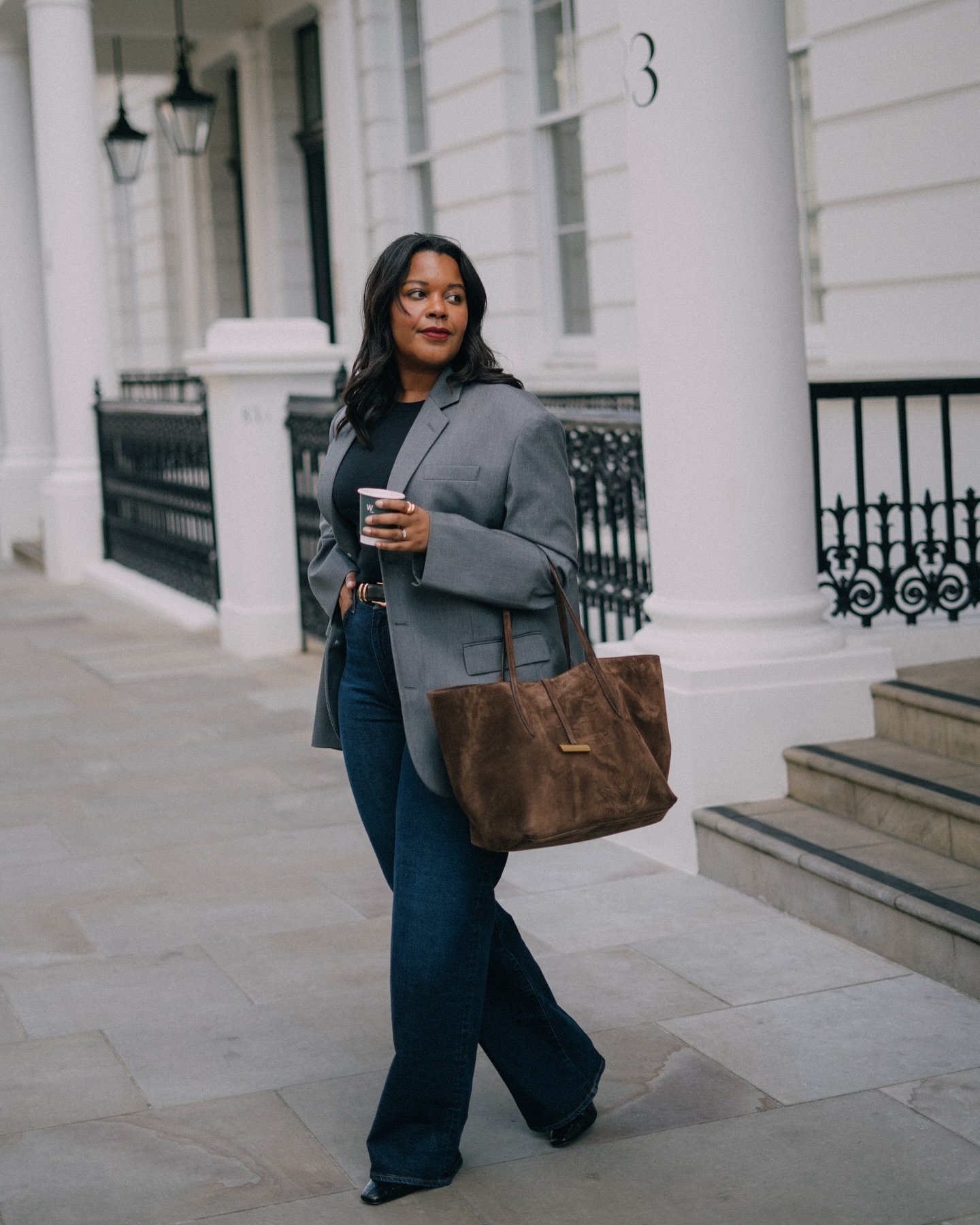 Mulher elegante com blazer cinza, jeans e bolsa marrom caminha segurando um café.