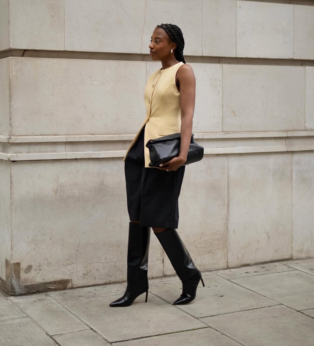 Pessoa vestindo colete bege e saia preta, segurando uma clutch, usando botas de cano alto. Um dos melhores looks de trabalho.