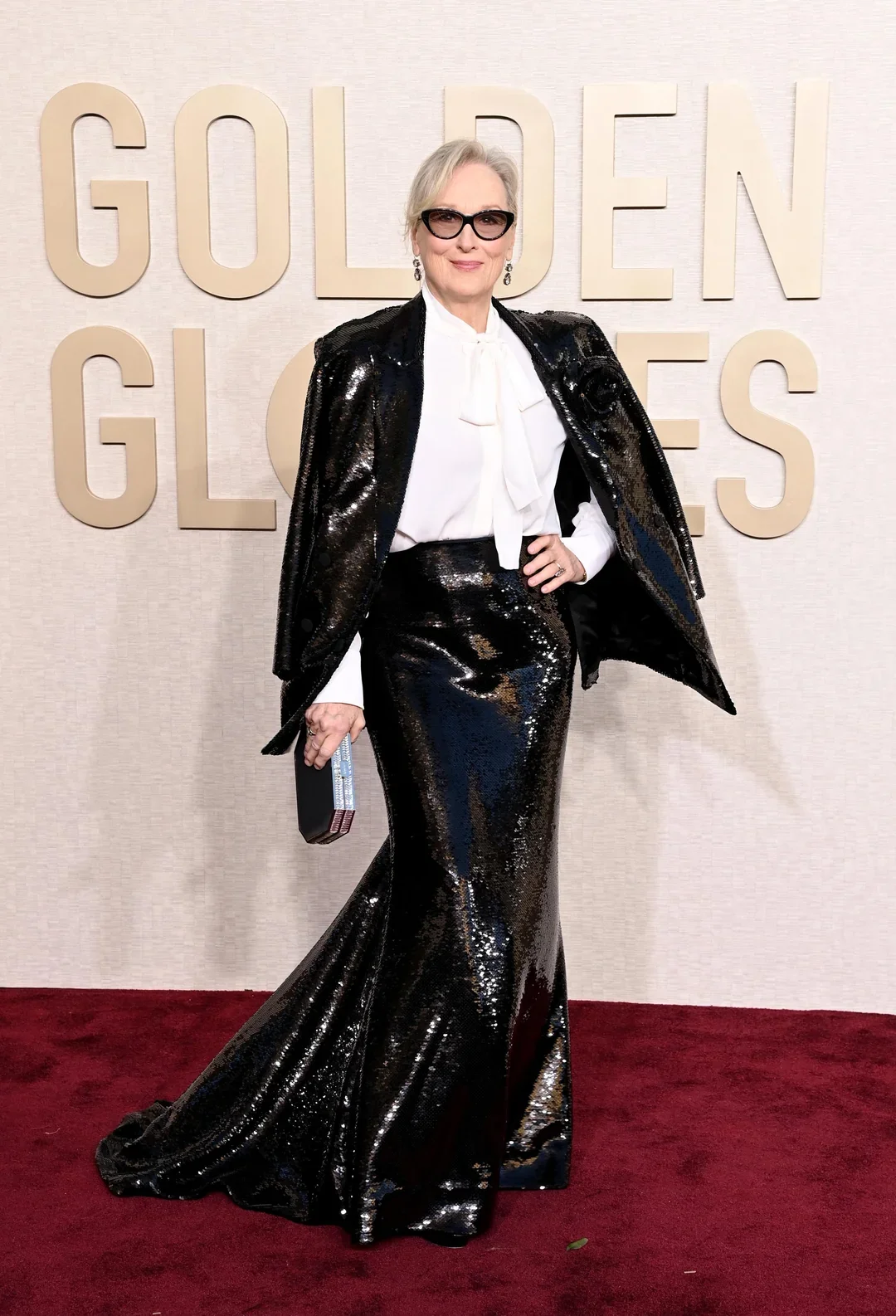 Pessoa elegante usando vestido preto brilhante e camisa branca com laço, segurando uma clutch, ao lado de logotipo de premiação.