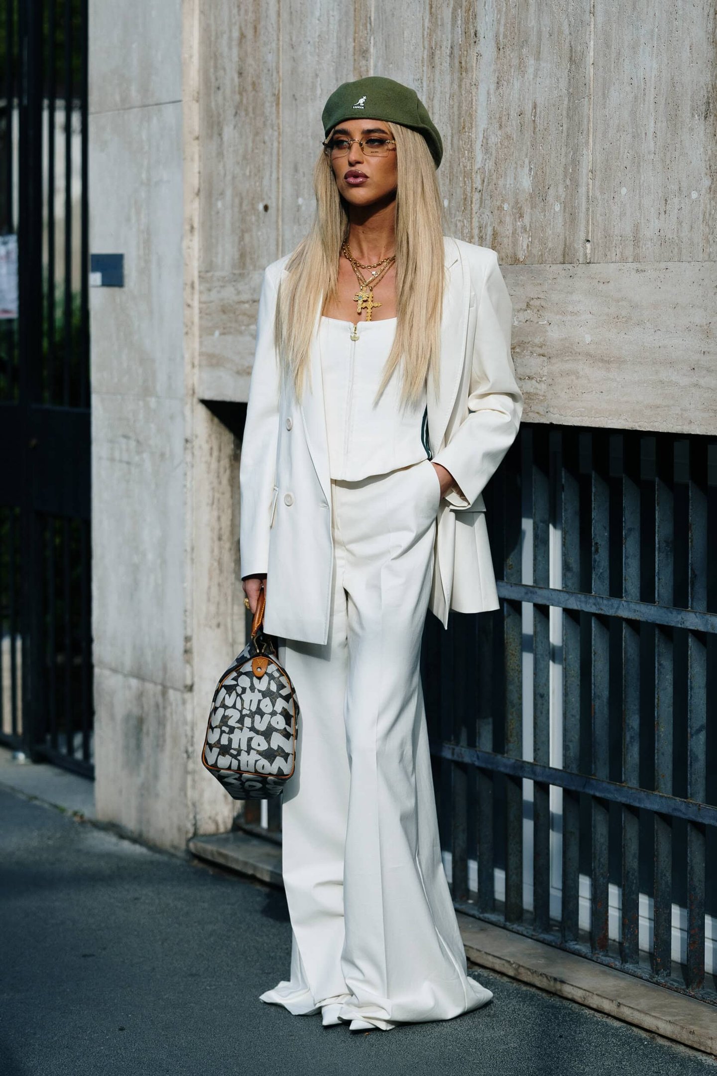 Mulher loira com boina verde, terno branco elegante e bolsa estampada. Acessórios dourados adicionam truques de estilo fáceis.