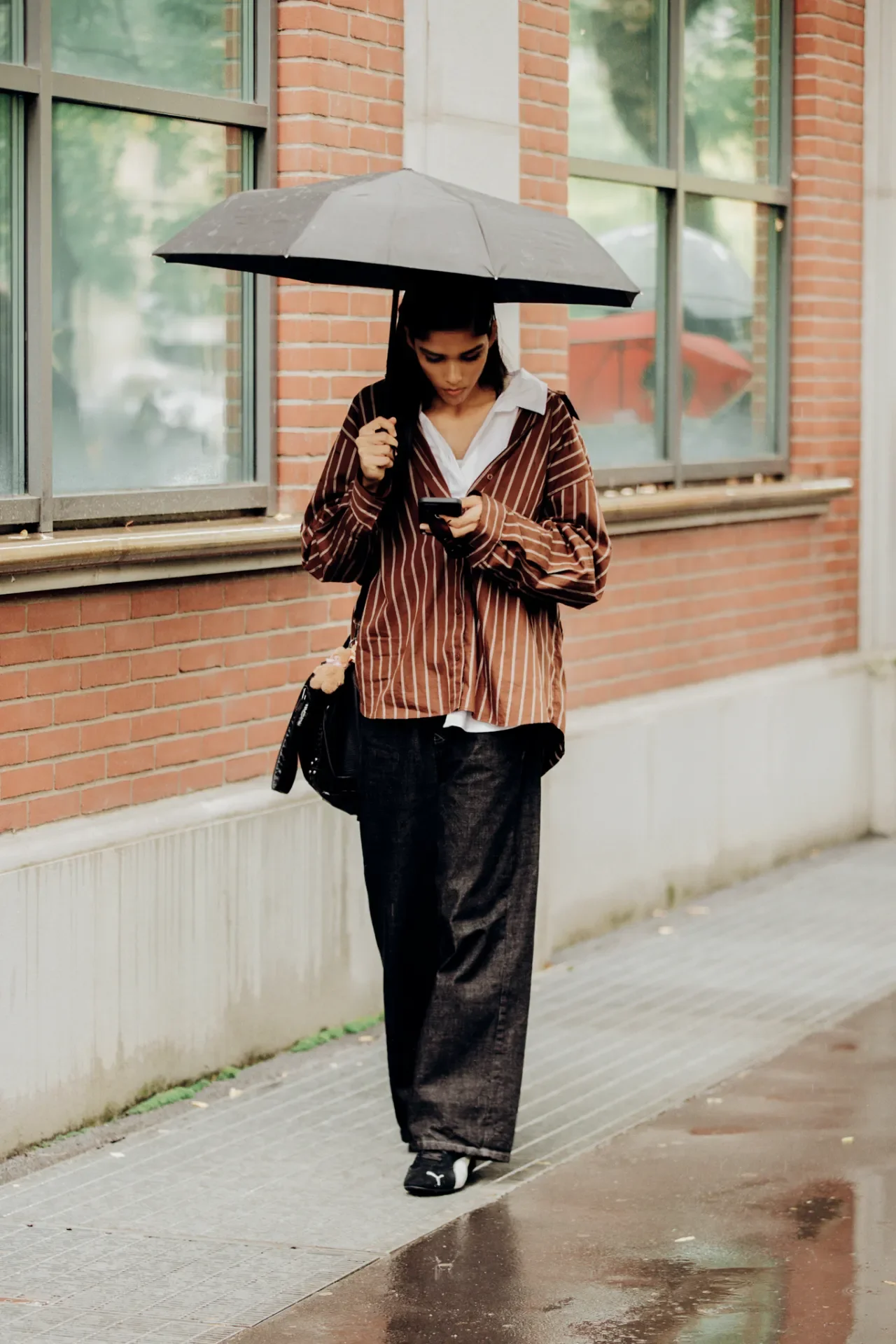 Usuária com guarda-chuva preto, camisa listrada e calça larga caminha sob chuva, representando truques de estilo fáceis e práticos.