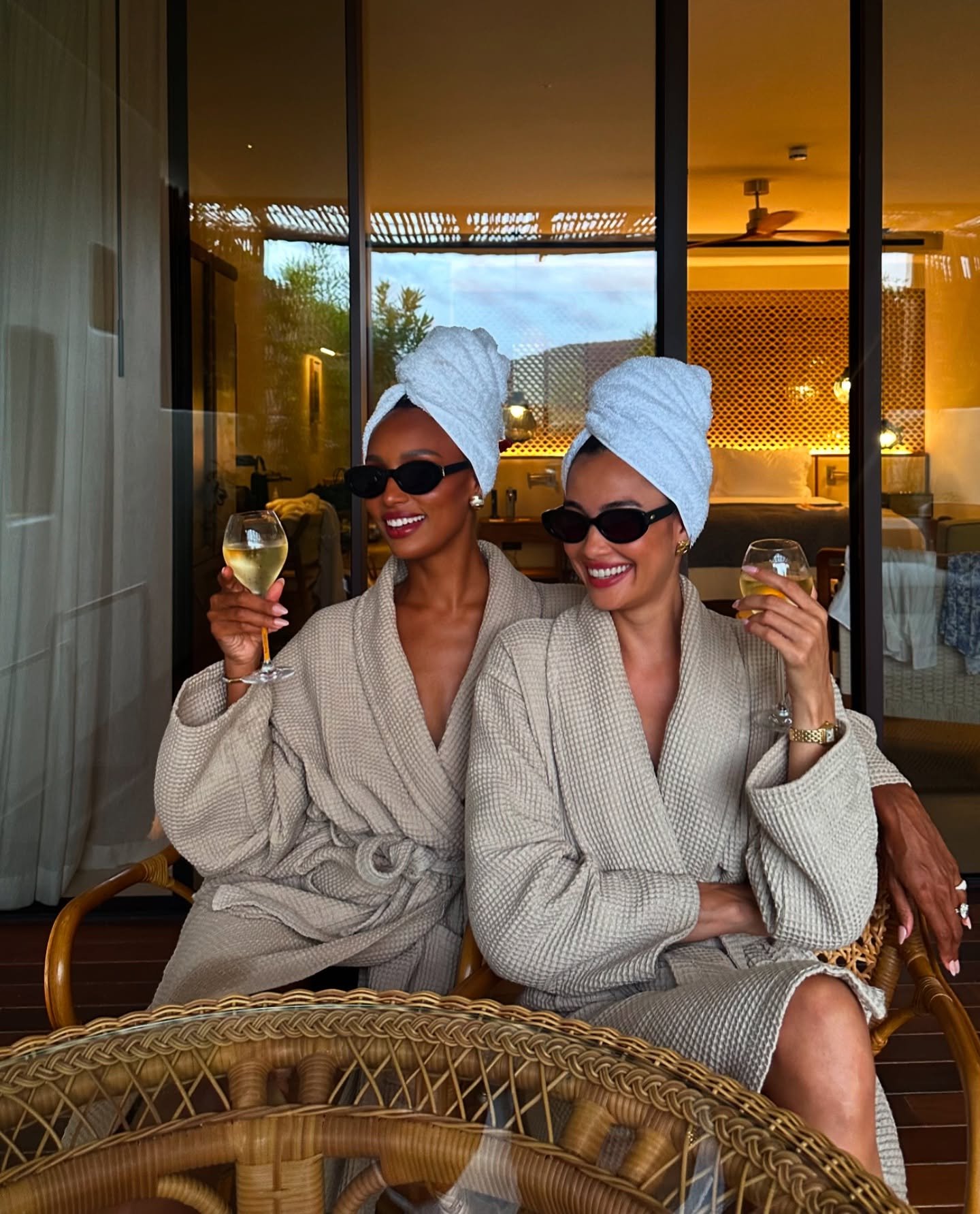 Duas mulheres sorrindo em roupões e toalhas na cabeça. Estão em uma varanda, segurando taças de vinho branco.