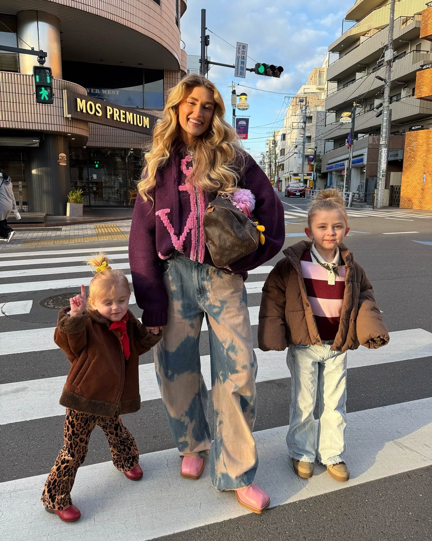 Pessoas atravessando rua em cidade, uma mulher com cabelo loiro e duas crianças ao lado, destacando laços familiares.