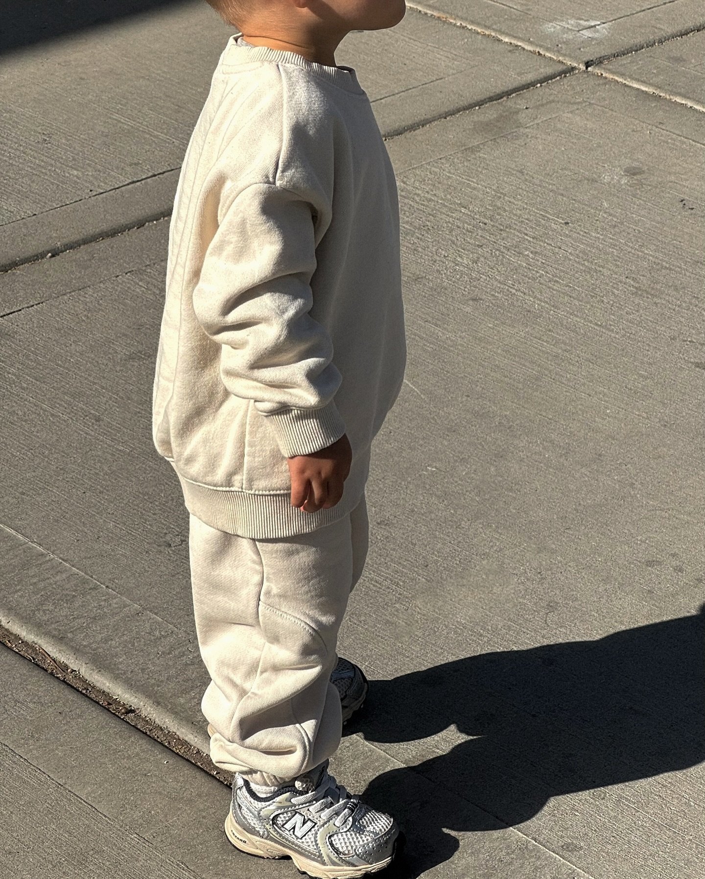 Criança de uniforme bege e tênis cinza em calçada ensolarada, projetando sombra no chão.