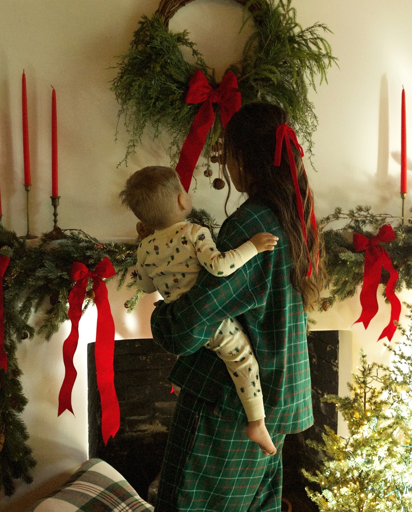 Mãe e criança em frente a uma lareira decorada para o Natal, exibindo laços vermelhos e velas acesas.