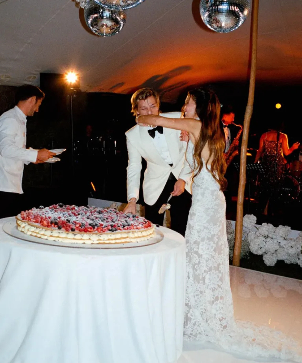 Casal elegante corta bolo em festa, com mulher usando vestido não tradicional repleto de rendas.