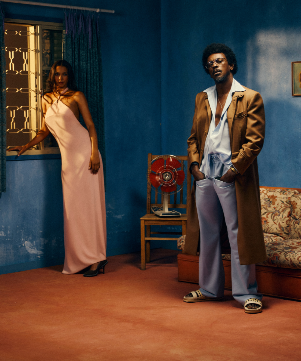 Uma mulher de vestido rosa e um homem de casaco marrom posam em um cômodo de estilo vintage para a campanha Tieta de Misci.