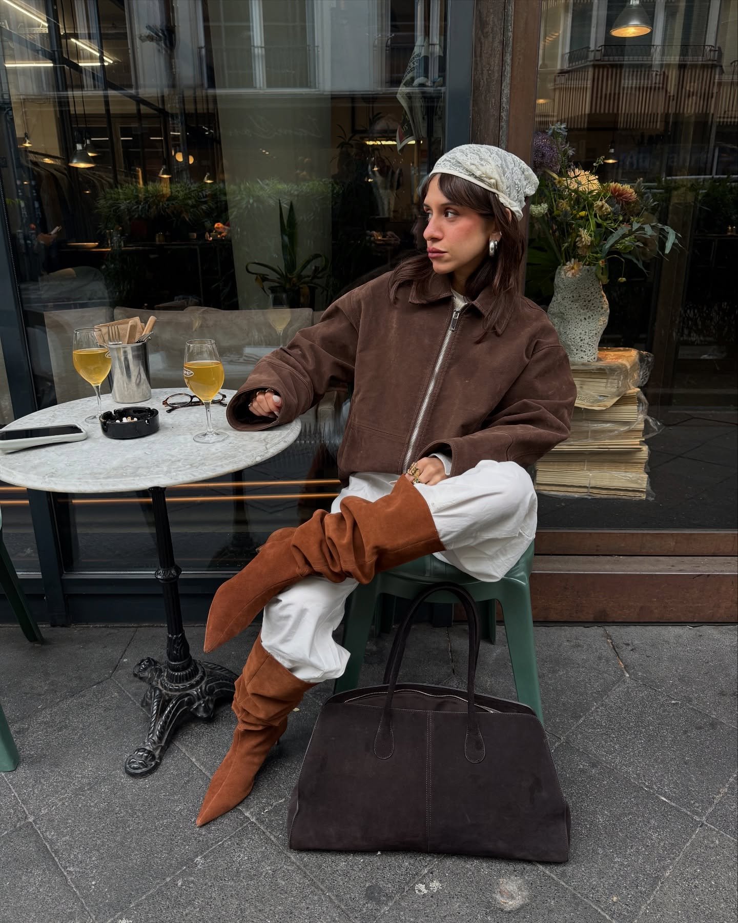 Pessoa sentada em café usa botas estilo boho marrons, calças brancas e jaqueta marrom. Bebidas sobre a mesa.