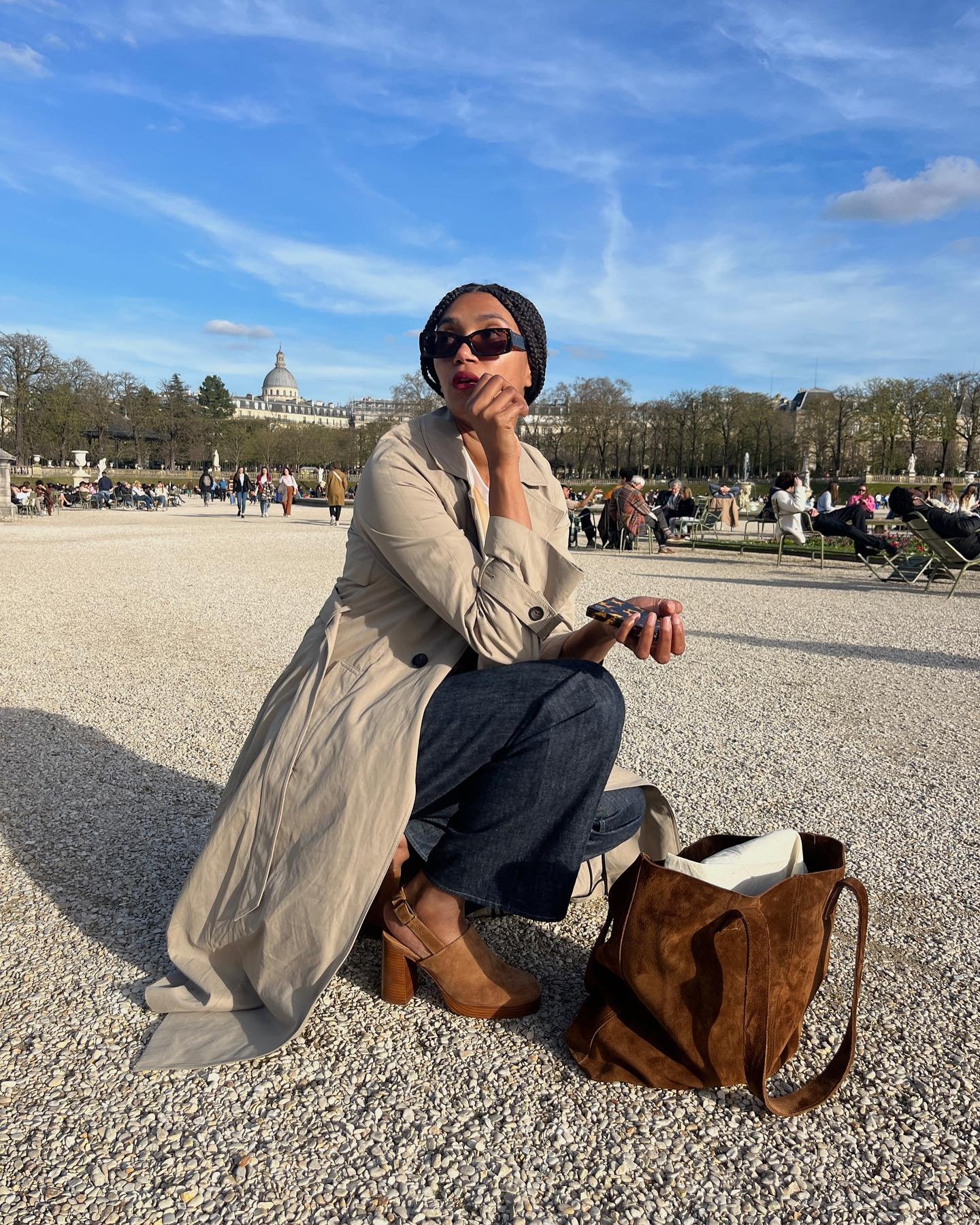 Pessoa com casaco bege e jeans, usa par de sapato boho marrom ao ar livre em um parque movimentado.