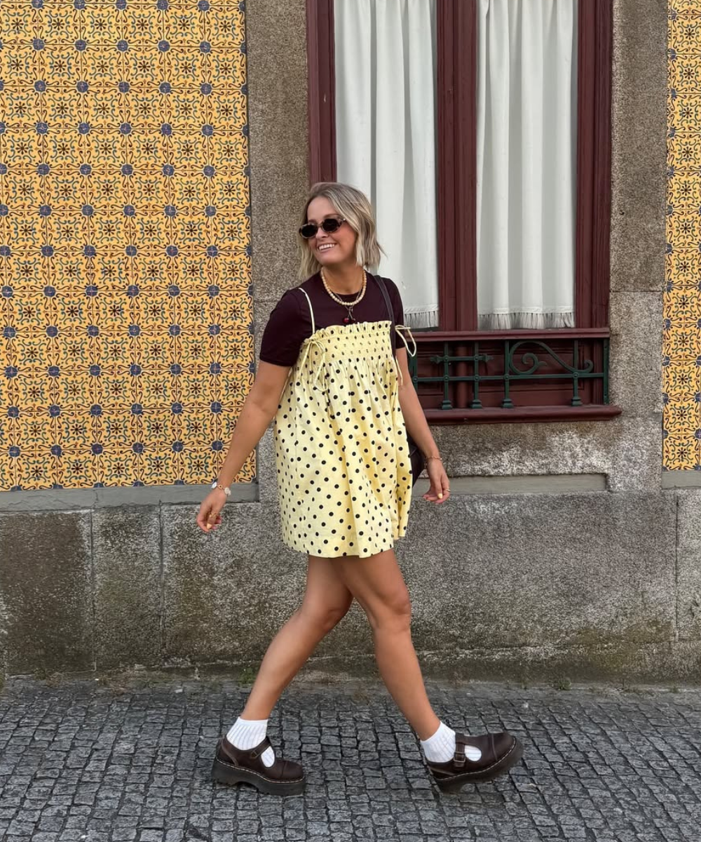 Pessoa com vestido leve de primavera em bolinhas, andando na calçada com parede de azulejos ao fundo.