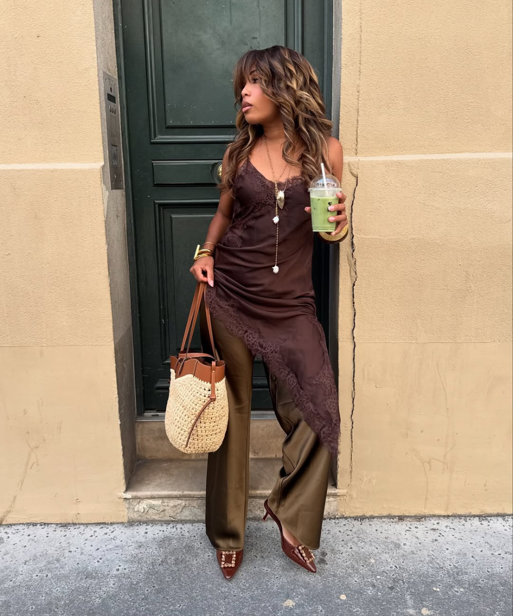 Pessoa em frente a uma porta verde, vestindo uma roupa marrom e segura uma bebida e bolsa.