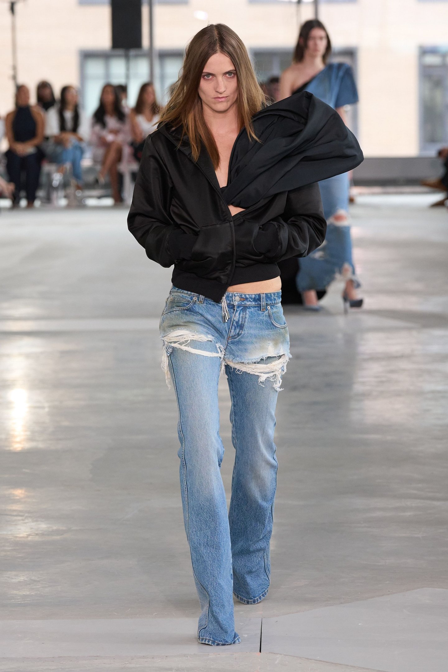 Modelo com jaqueta preta e jeans rasgado desfila na New York Fashion Week SS26. Público ao fundo.