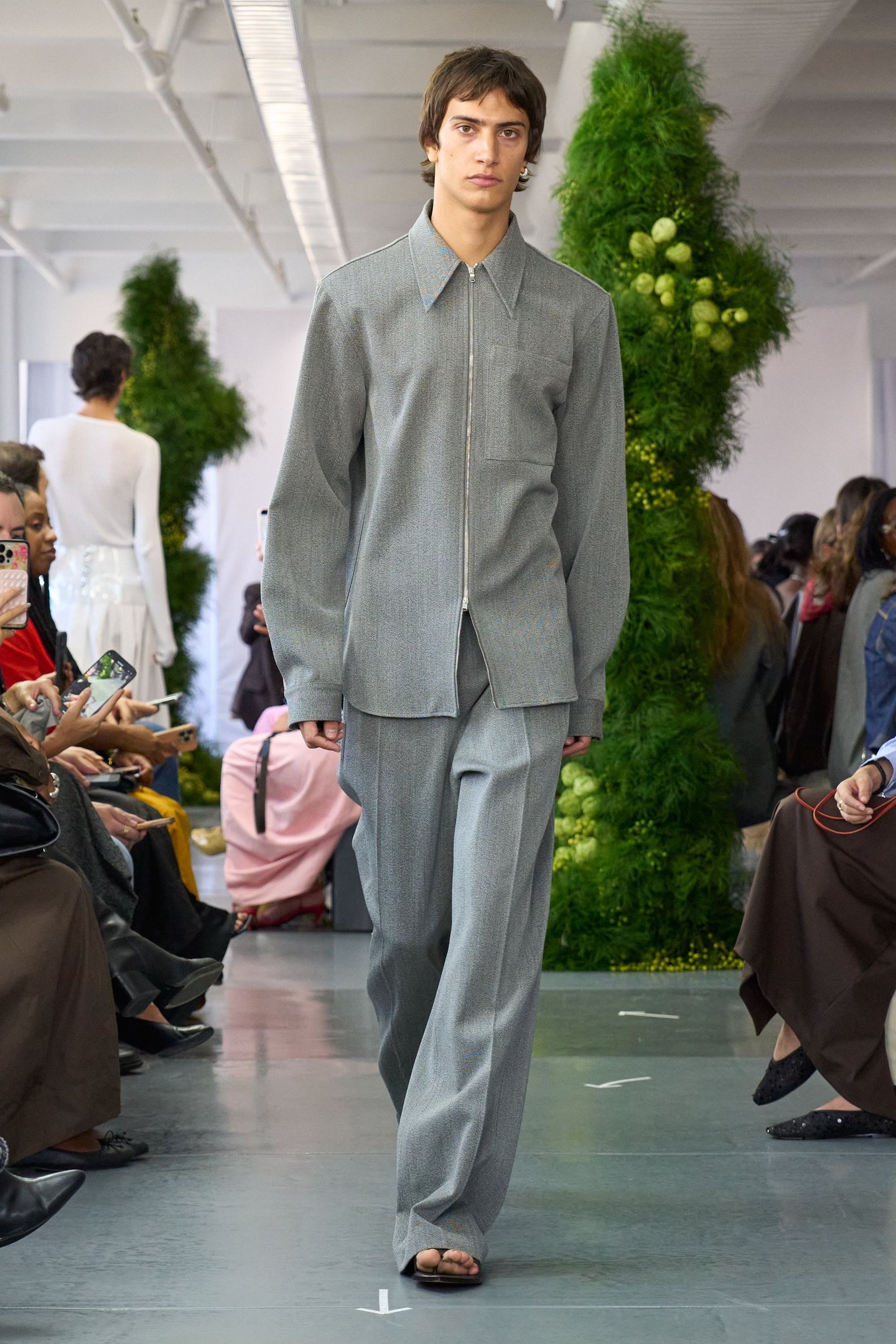 Desfile na New York Fashion Week SS26 com modelo vestindo conjunto cinza e público ao fundo.