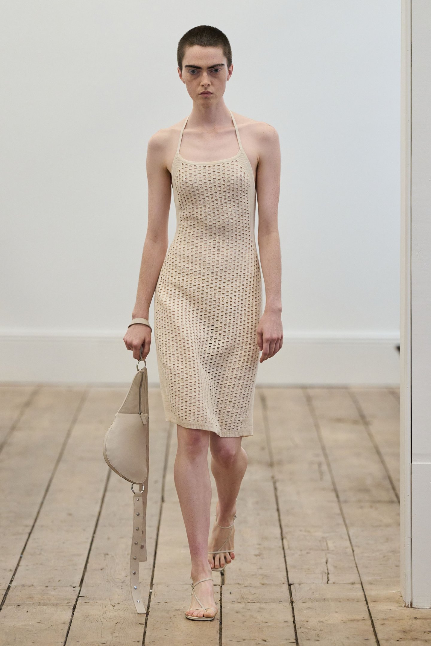 Modelo em vestido bege de crochê desfila na New York Fashion Week SS26, segurando bolsa bege.