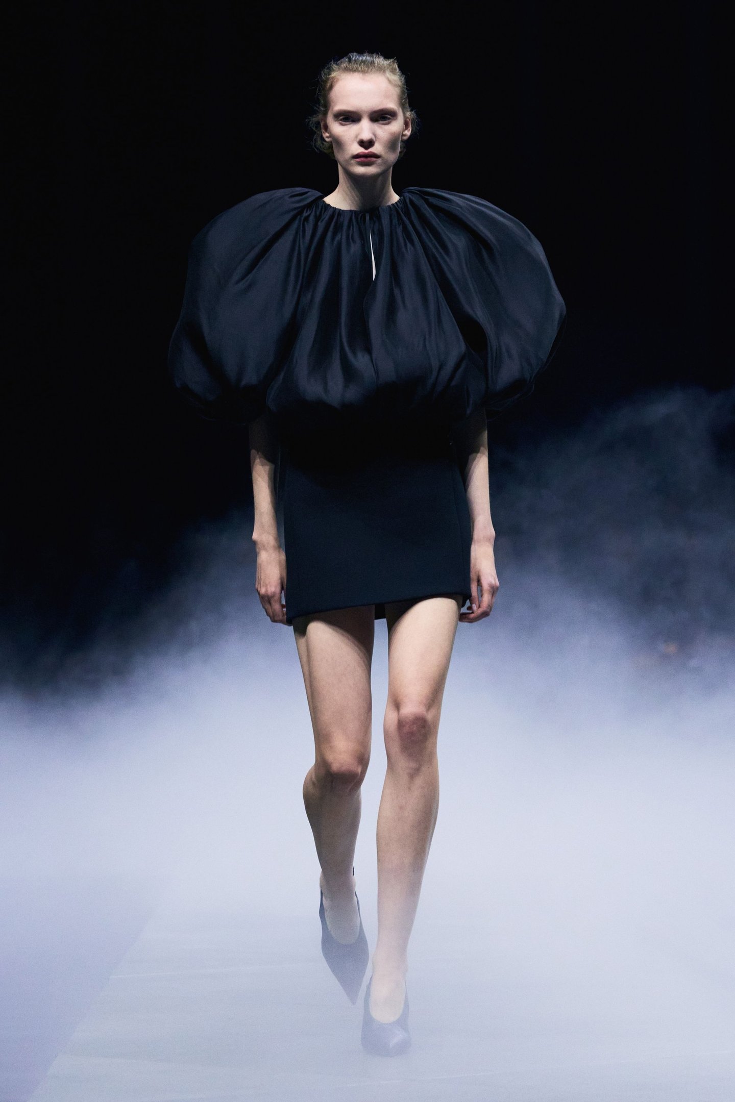 Modelo com vestido preto volumoso na New York Fashion Week SS26. Passarela com fundo escuro e névoa discreta.