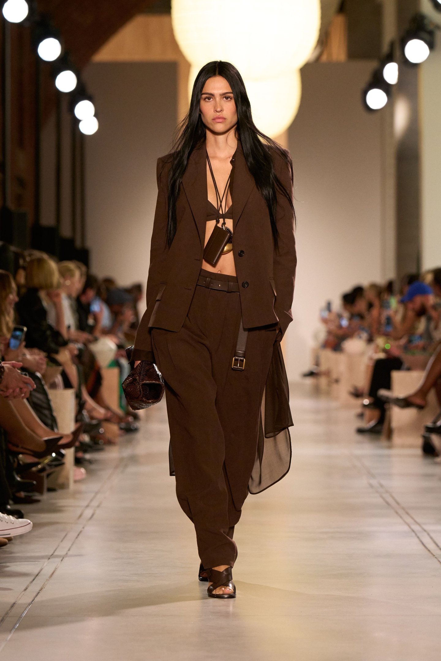 Modelo desfila com conjunto marrom na New York Fashion Week SS26, em uma passarela cercada por público atento.