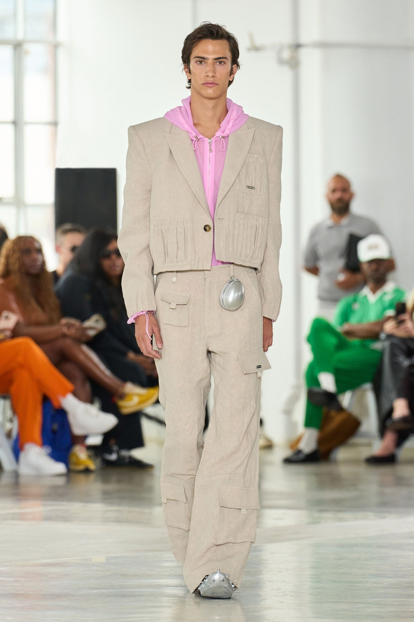 Modelo desfila na New York Fashion Week SS26 usando conjunto bege com detalhes plissados e moletom rosa.