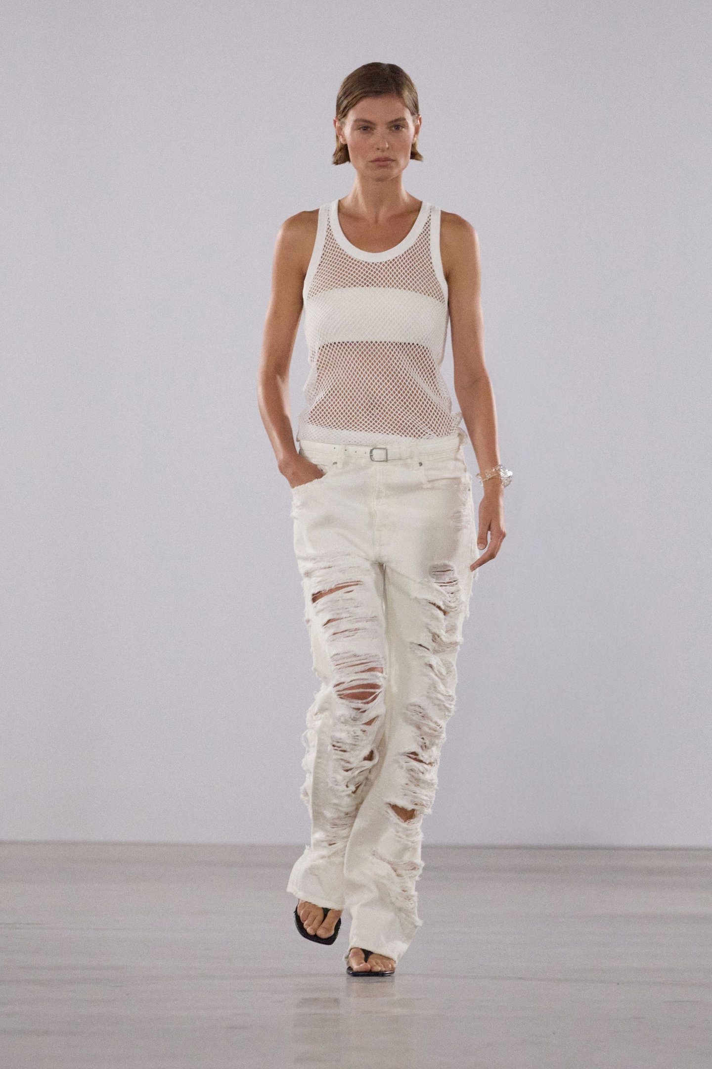 Modelo desfila na New York Fashion Week SS26 usando blusa de tela branca e calça rasgada.