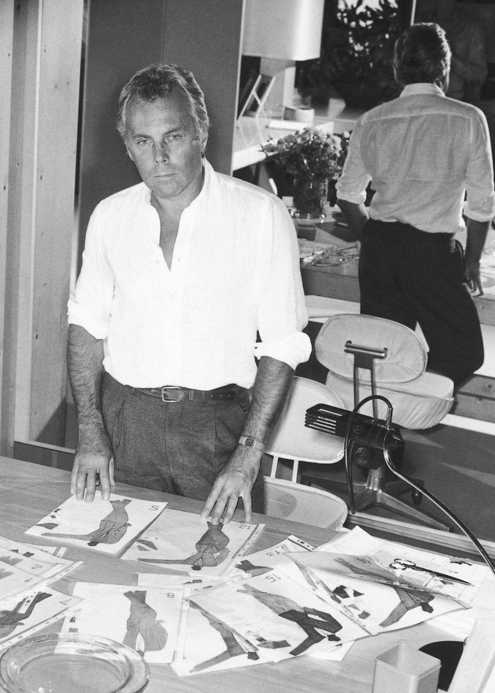 Homem em escritório revisa esboços de moda sobre mesa. O estilo pode ser associado ao design de Giorgio Armani.
