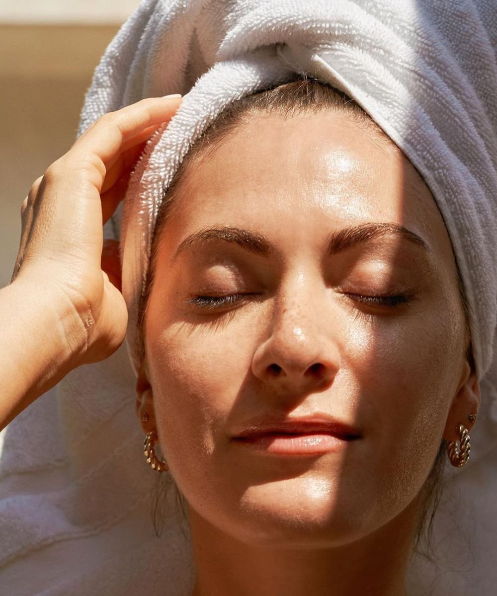 Mulher relaxada com toalha na cabeça, olhos fechados, pele brilhante como após uso de água micelar.