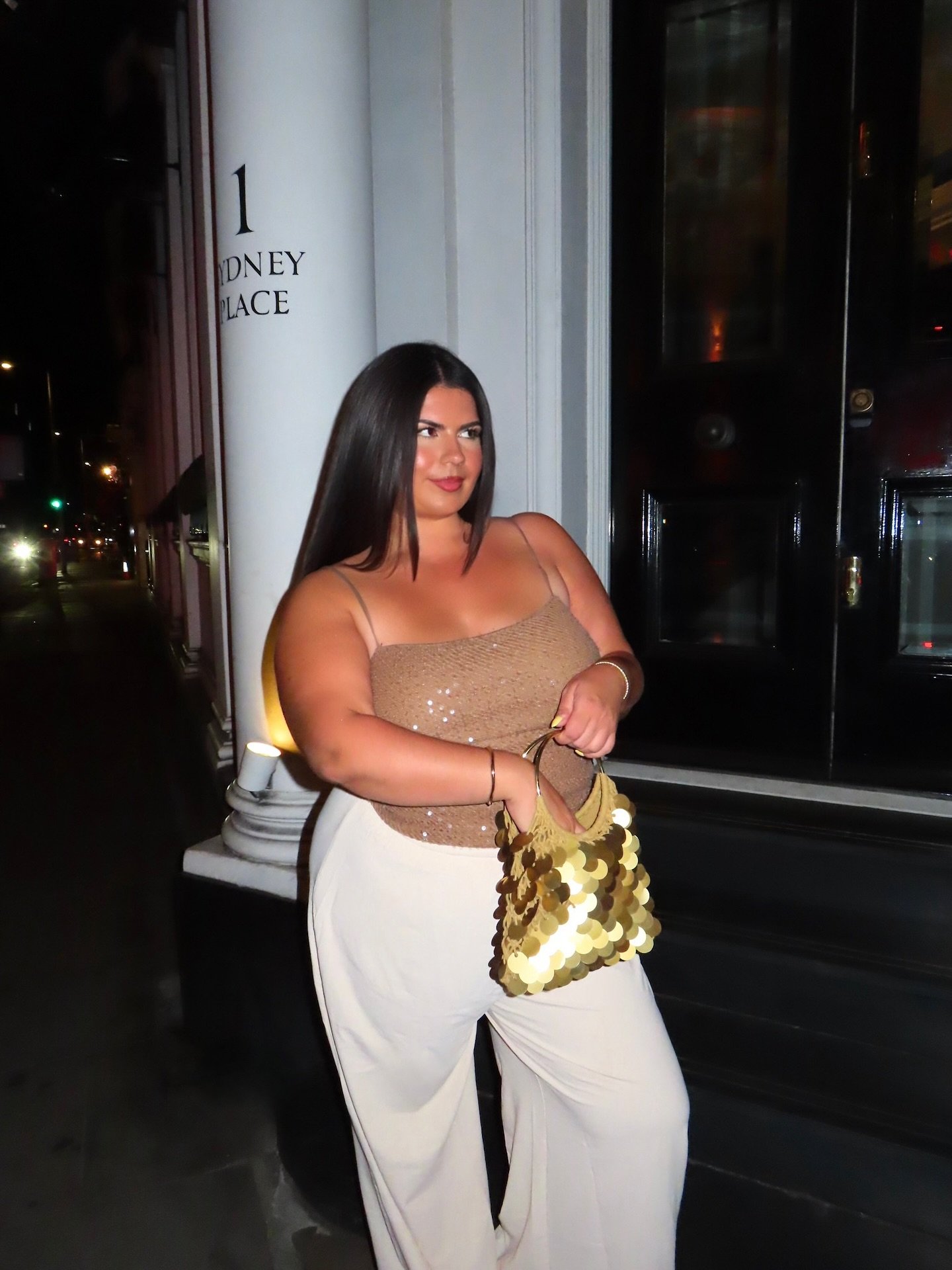 Pessoa com bolsa dourada e top brilhante posa à noite em local elegante, ideal para festas.