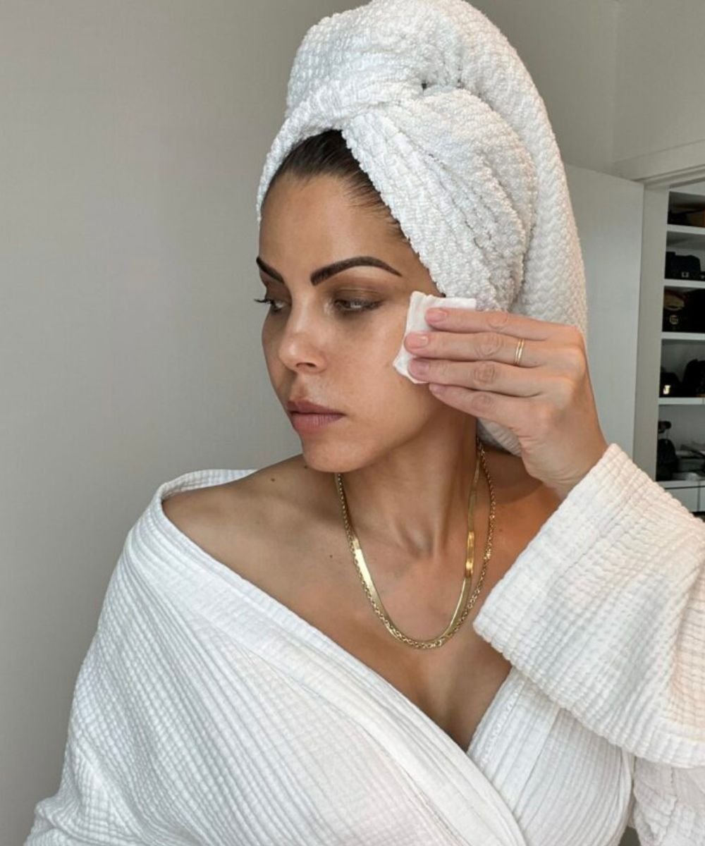 Mulher fazendo limpeza facial com algodão embebido em água micelar, toalha na cabeça e roupão branco.