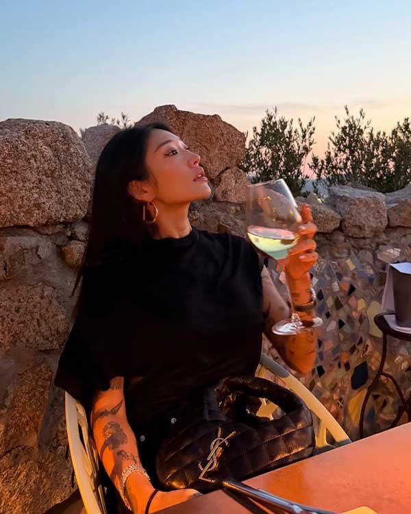 Mulher tatuada segura taça de vinho ao pôr do sol, bolsa Icarino da Saint Laurent no colo.