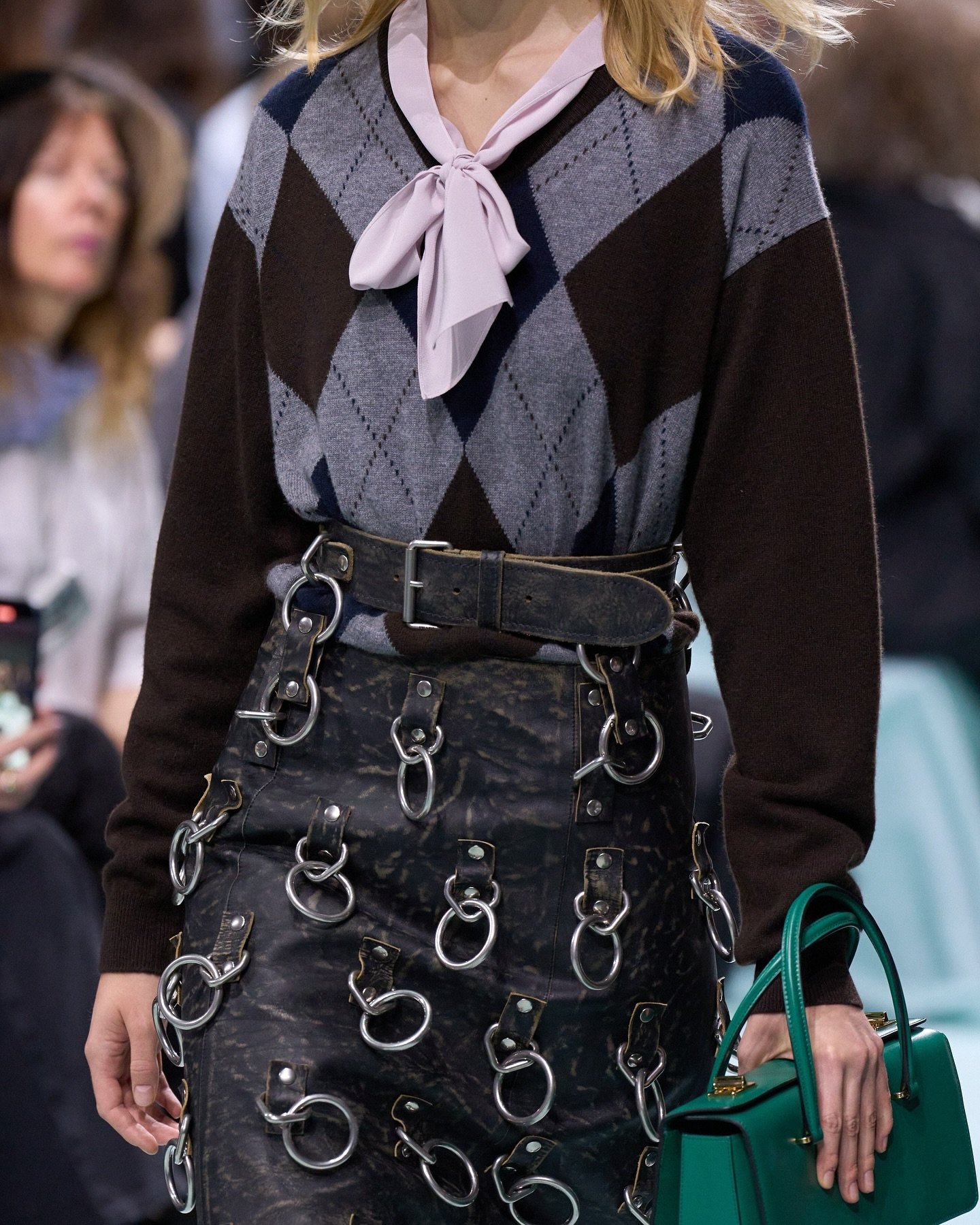 Suéter argyle e saia com apliques circulares, refletindo a moda dos piercings, em desfile de moda.