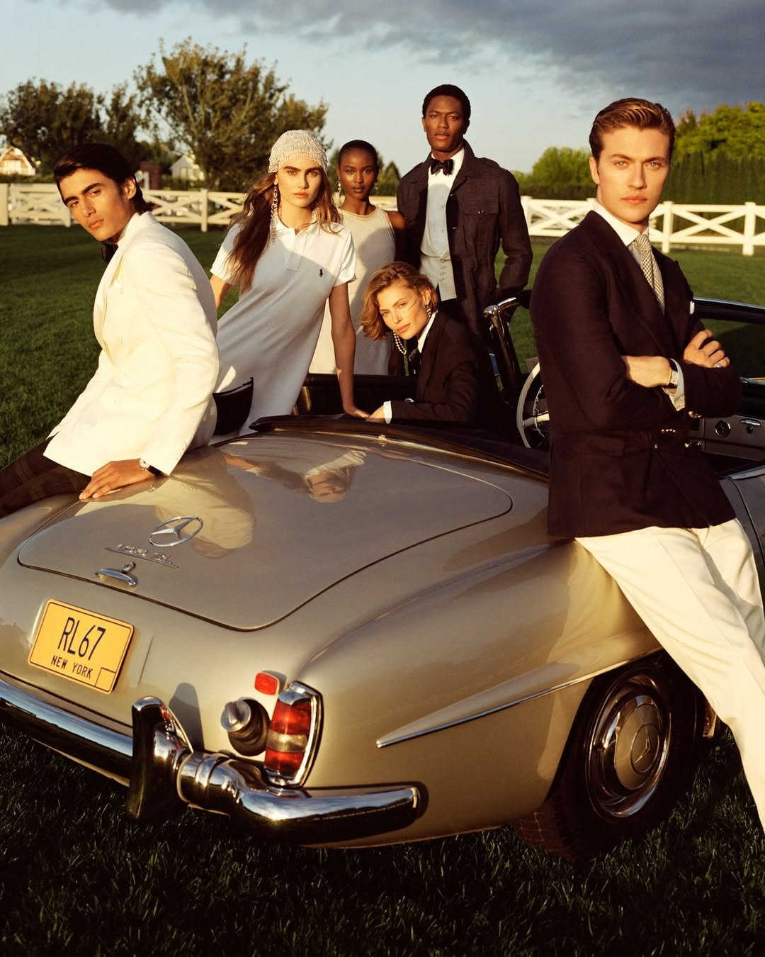 Grupo de jovens em roupas elegantes ao lado de um carro clássico em cenário ao ar livre, com estilo preppy.