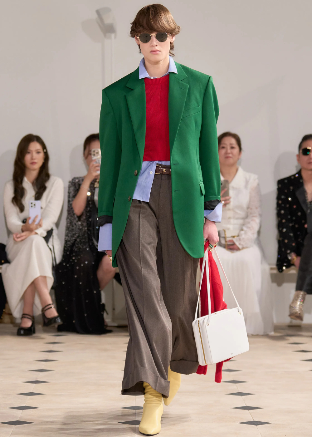 Modelo na passarela com blazer verde, suéter vermelho e calça larga, em um estilo preppy contemporâneo.