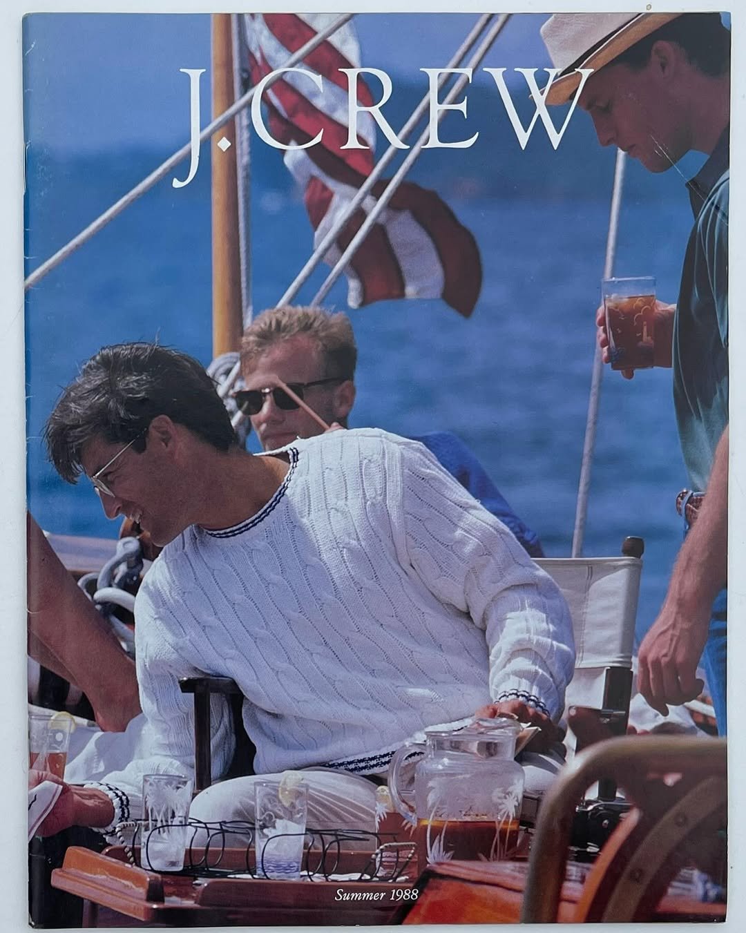 Homens em um barco, usando roupas estilo preppy, desfrutando de uma bebida. Ao fundo, uma bandeira e o mar.