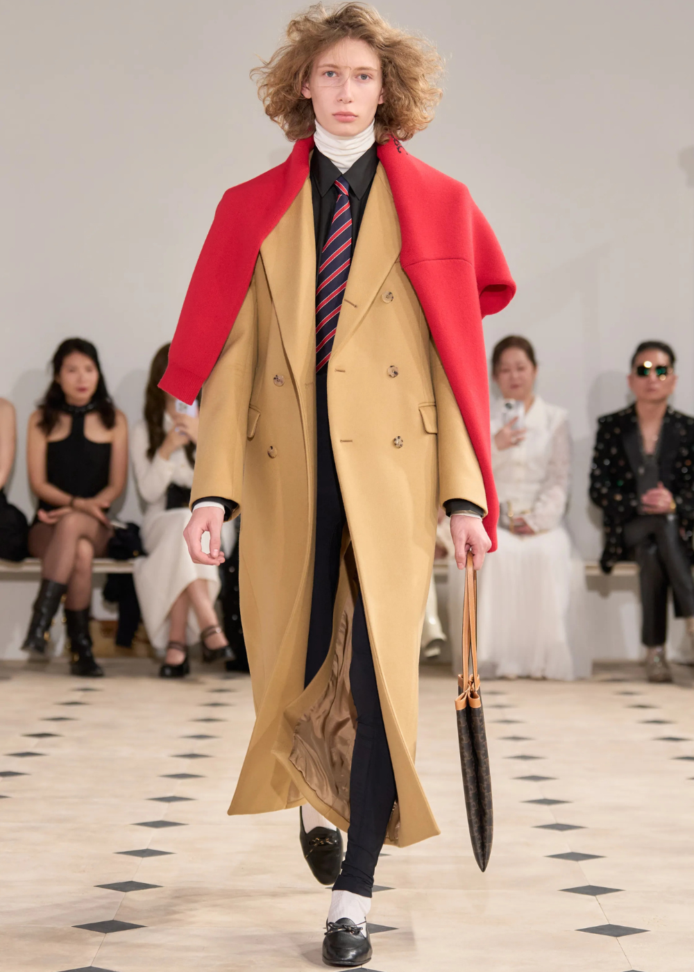 Modelo na passarela com visual preppy, usando sobretudo bege, capa vermelha, gravata listrada e bolsa de couro.
