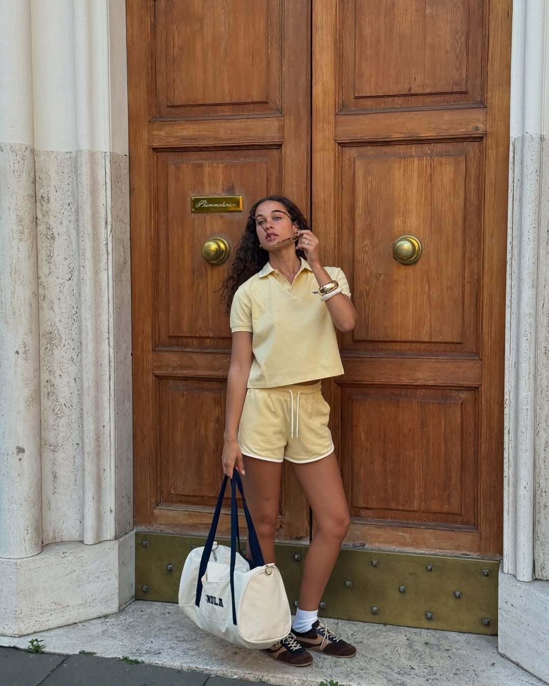Jovem em frente à porta de madeira, veste conjunto amarelo e segura bolsa, estilo esportivo preppy.
