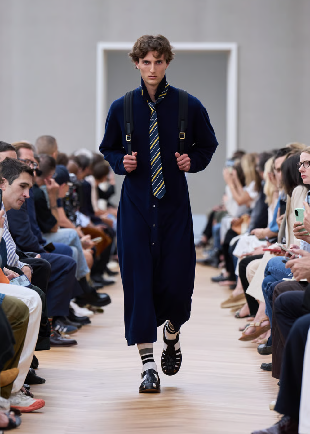 Desfile de moda masculina com traje azul escuro, gravata listrada e estilo preppy. Público nas laterais.
