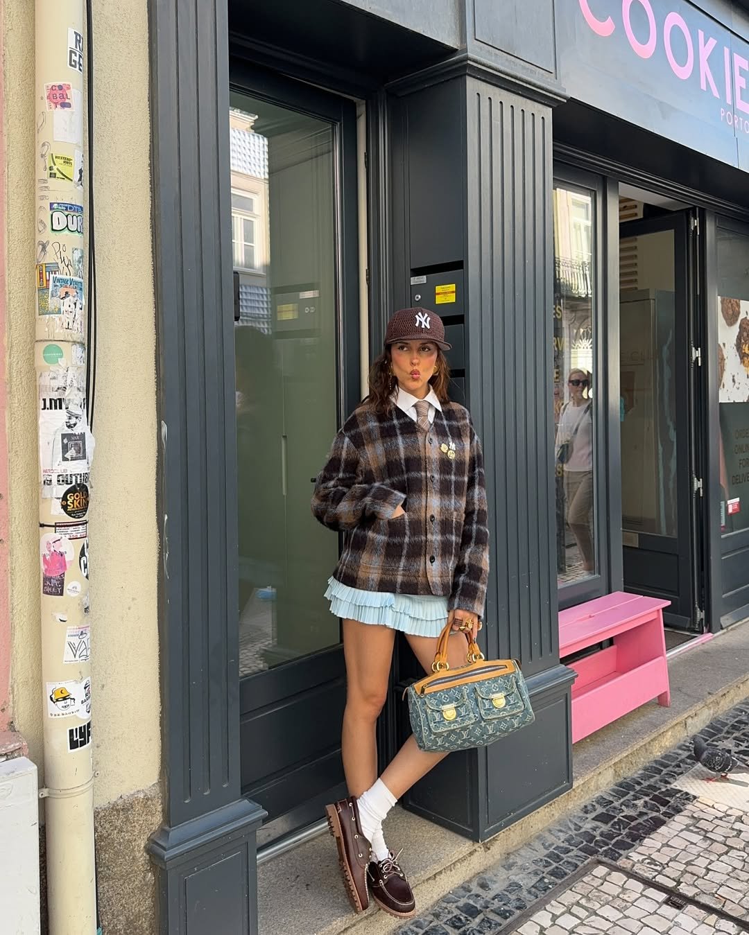 Pessoa com visual preppy usa casaco xadrez e saia plissada, enquanto segura uma bolsa. Ela está na calçada.