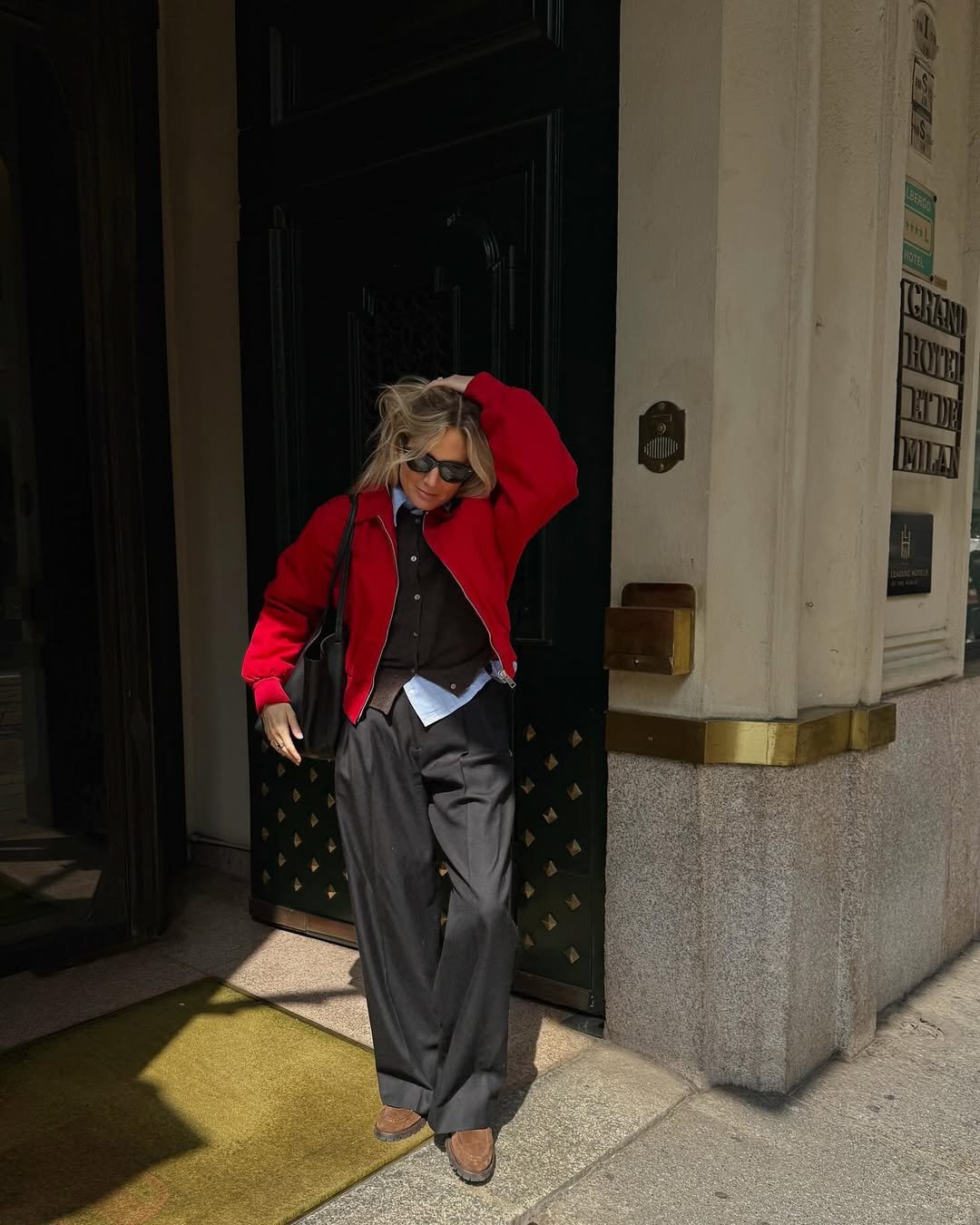 Pessoa com casaco vermelho elegante, óculos escuros e calças largas, exibe um estilo preppy em frente a prédio.